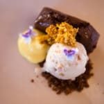 A dessert of two scoops of icecream, one pale yellow and the other white with honeycomb on the top, the white scoop of icecream sits on chocolate crumble and behind both scoops of icecream is a rectangular piece of gingerbread cake. The dessert image is set justified to the top right of the imageon an ombre pale purple and orange background