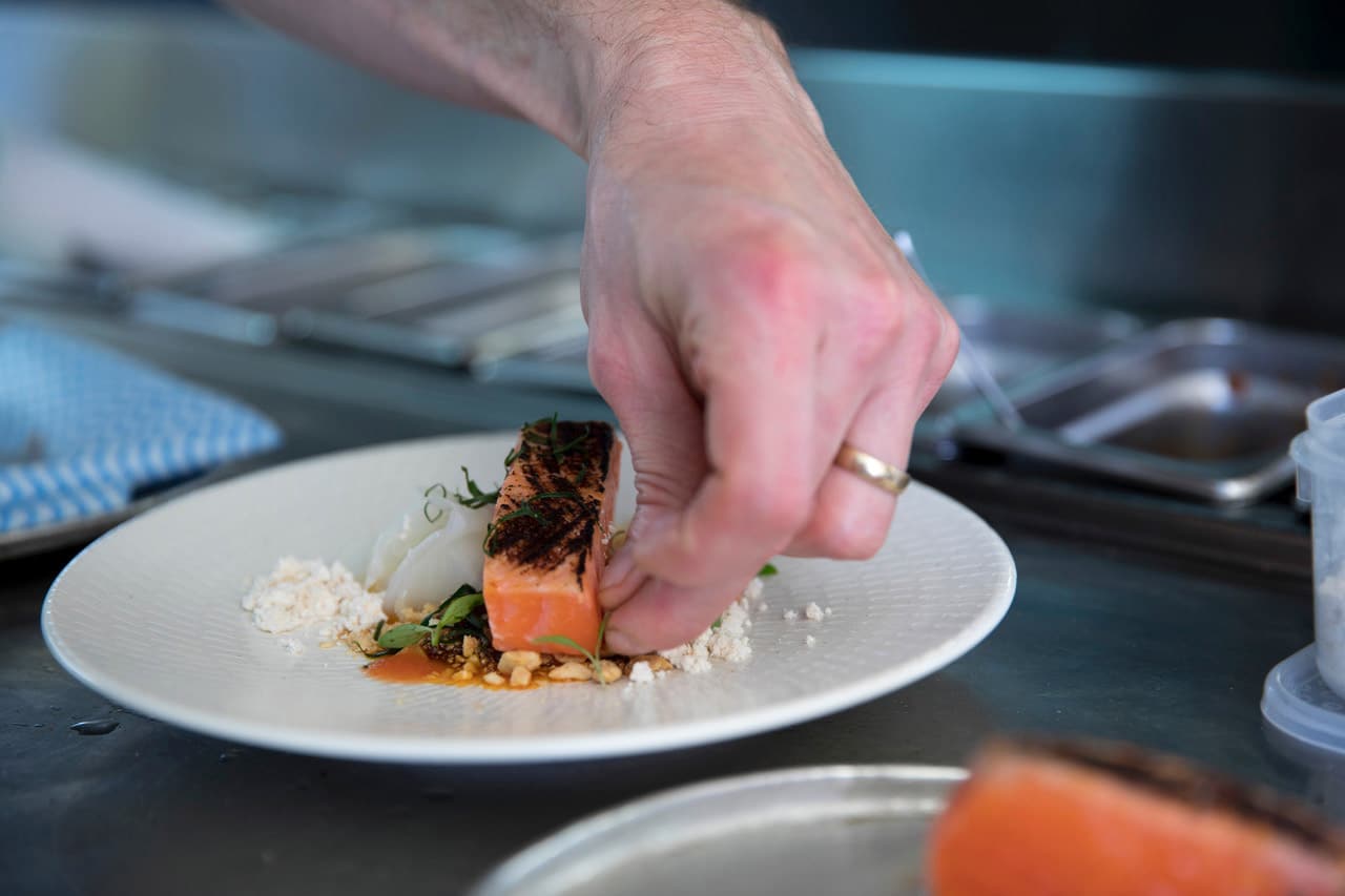A chef's hand with gold ring places a micro green garnish on a white plate containing blackened salmon, an orange sauce and some accompanying food items that are hard to say exactly what they are, an opaque vegetable or perhaps a softened rice noodle, some crumbled cheese or perhaps cauliflower and what appears to be some pinenuts or chopped hazelnuts. In the background a chef's preparation bench is blurred but still able to be distinguished