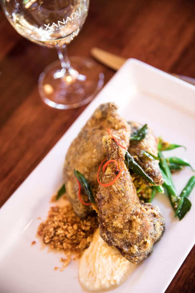 A white oblong-shaped plate with a bevelled edge takes up two thirds of the image and is on a forty five degree angle. A wine glass stem and a dark wooden table form the border of this plate in the top left hand side of the image. The plate containsexample of lunch at Xanadu wines achar spatchcock green beans paneer peanut praline